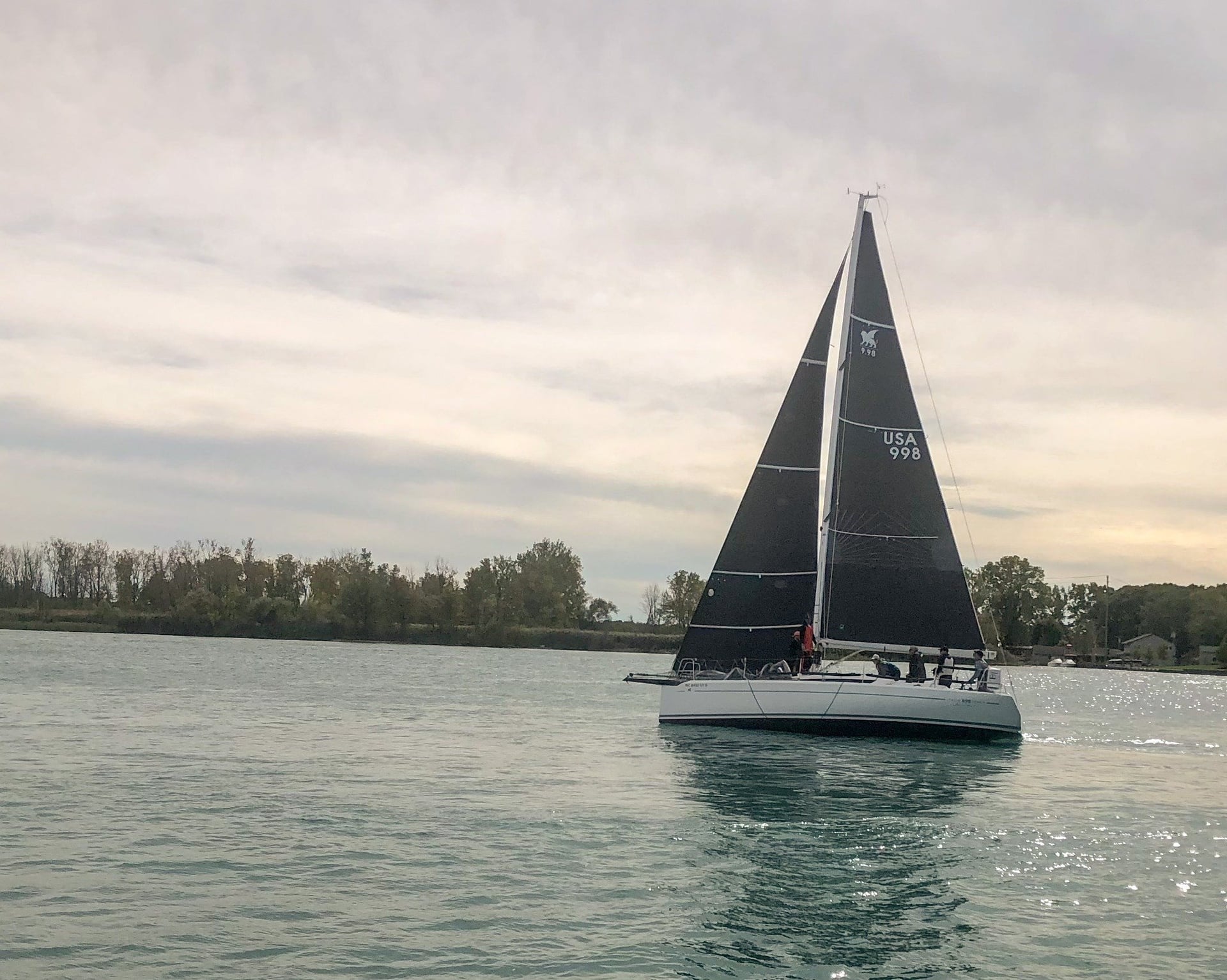Sailboat with black sails and crew along a tree-lined shore